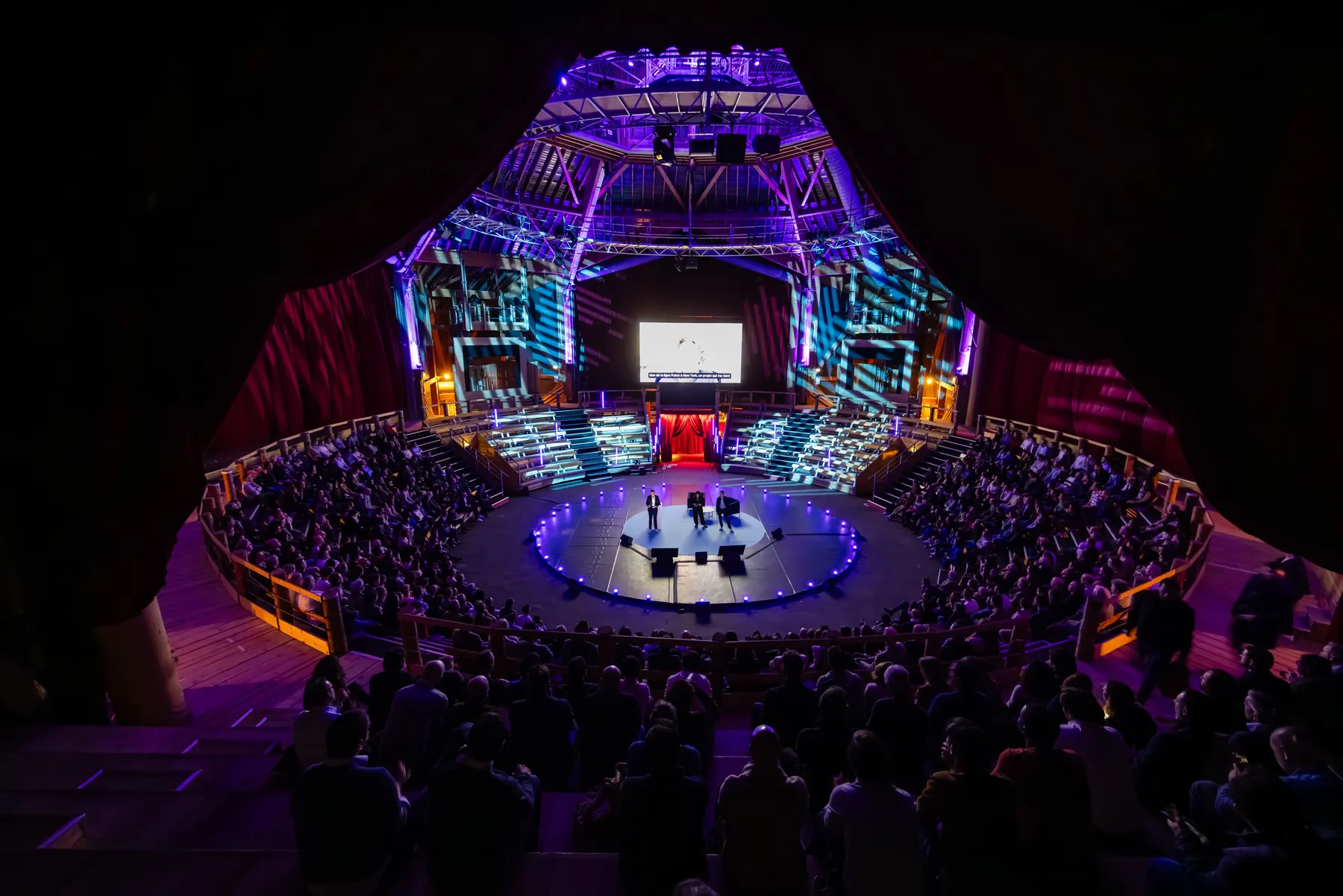 Vue en hauteur d’une scène circulaire entourée de gradins, sous des lumières violettes