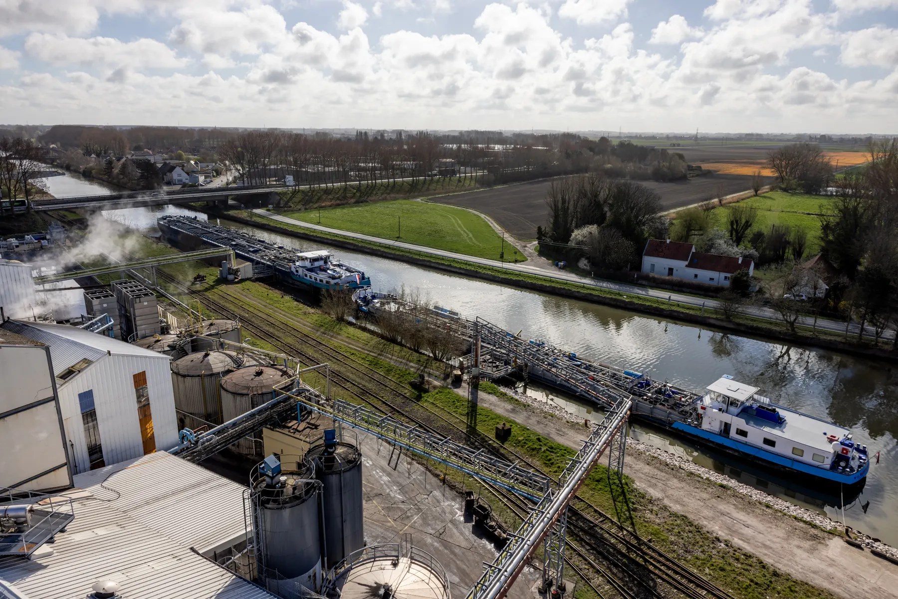 Vue aérienne d’un site industriel au bord d’un canal : cuves, voies ferrées et péniches