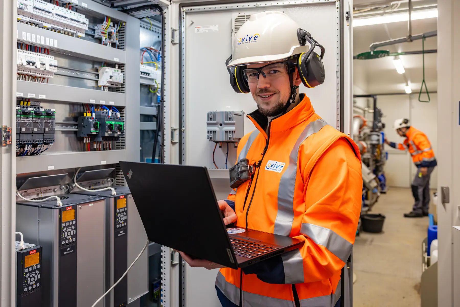 Technicien en casque et veste haute visibilité, ordinateur en main devant des armoires électriques